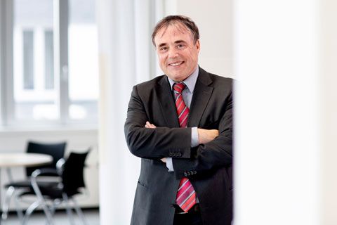 Ein lächelnder älterer Mann im Anzug mit dunklen Haaren steht vor einem Besprechungsraum